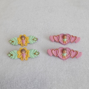 May include: Four small decorative hair clips. Two clips feature a yellow flower with a small doll in a pink dress, surrounded by green leaves and heart accents. The other two clips have a pink heart design with a doll in a green dress.