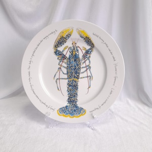 May include: White ceramic plate featuring a detailed illustration of a blue and yellow lobster. The lobster design is centered on the plate, with text around the rim. The plate is displayed on a clear stand.
