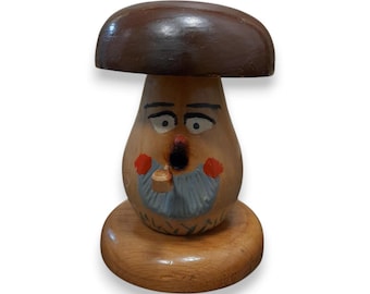 Puede incluir: Una caprichosa figura de hongo de madera con un sombrero marrón y una cara tallada en el tallo. La cara tiene ojos pintados, una nariz roja y una barba azul. La figura está montada sobre una base redonda de madera.