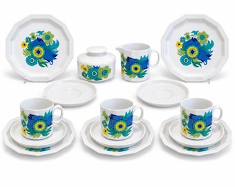 Może przedstawiać: Biały ceramiczny zestaw do herbaty z żywym kwiatowym wzorem w odcieniach niebieskiego, zielonego i żółtego. Zestaw zawiera trzy filiżanki ze spodkami, dwa większe talerze, cukiernicę z pokrywką oraz dzbanek na mleko, wszystkie z ośmiokątnym brzegiem.