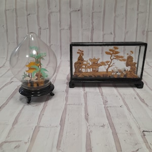 Può includere: Due sculture in sughero racchiuse in vetro con scene di ispirazione asiatica. Una è una cupola rotonda con una pagoda e alberi. L'altra è una scatola rettangolare con una pagoda, alberi e un ponte.