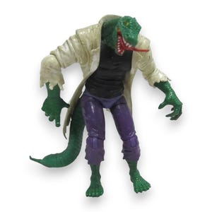 Könnte beinhalten: Eine grüne, echsenartige Actionfigur mit weißem Mantel, schwarzem Hemd und lila Hose. Die Figur hat einen langen Schwanz und ist mit ausgestreckten Armen posiert. Die Echse hat eine rote Zunge und steht auf weißem Hintergrund.