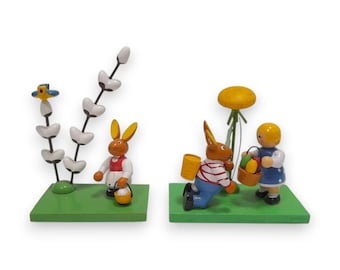 Op de afbeelding: Twee houten paasbeeldjes op groene bases. Eén toont een konijn met een mandje bij een plant. De andere toont een konijn dat knielt met een mandje en een kind dat een mandje met eieren vasthoudt bij een bloem.