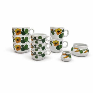 Pode incluir: Um conjunto de loiça de cerâmica branca com um padrão repetitivo de flores amarelas e cor de laranja com folhas verdes. O conjunto inclui sete canecas, uma pequena leiteira e uma taça pequena, todas decoradas com o mesmo motivo floral.