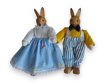 Peut inclure: Deux figurines de lapin en bois. L'un porte une blouse blanche et une jupe à carreaux bleus avec des accents colorés. L'autre porte une chemise jaune, une salopette rayée bleue et blanche et un nœud papillon noir. Les deux ont des têtes en bois et de longues oreilles.