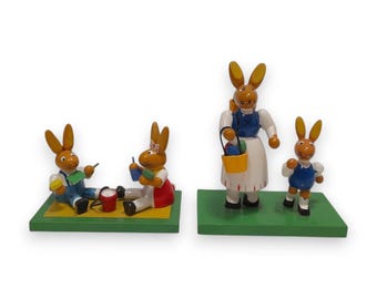 Peut inclure: Figurines de lapins de Pâques en bois. Deux lapins peignent des œufs, tandis qu'une lapine tient un panier d'œufs avec un petit lapin à côté d'elle. Les figurines sont sur des socles verts.