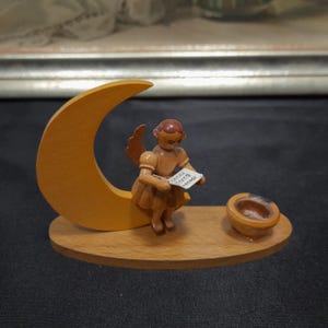 Könnte beinhalten: Eine Holzengelfigur, die auf einem Halbmond sitzt, mit einer kleinen Holzschale. Der Engel hält ein kleines Stück Papier mit dem Text "Gruss aus dem Erzgebirge" in der Hand.