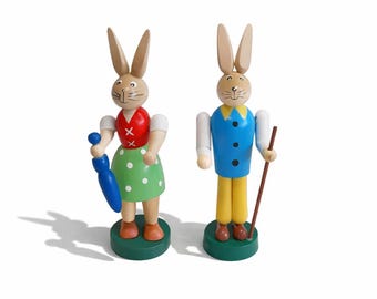 Peut inclure: Deux figurines de lapin en bois. L'un porte un gilet rouge, des manches blanches et une jupe verte à pois, tenant un parapluie bleu. L'autre porte un gilet bleu, un pantalon jaune et tient un bâton de marche marron. Les deux sont sur des socles verts.
