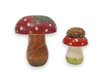 May include: Two decorative mushroom figurines. The larger mushroom has a red cap with white spots and a natural wood grain stem with green painted grass at the base. The smaller mushroom features a brown textured cap, a red ring, and a white stem.