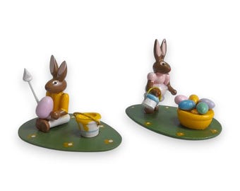 May include: Two wooden Easter bunny figurines. One bunny holds a spear and a pink egg, the other carries a paint bucket and stands near a bowl of colorful eggs. Both are on green bases.