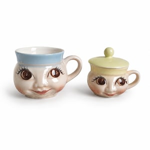 Peut inclure: Un mug en céramique avec un bord bleu clair et un petit récipient en céramique avec un couvercle jaune pâle. Les deux pièces présentent un visage moulé avec des yeux marron, de longs cils et des joues roses, avec une anse sur le côté droit.