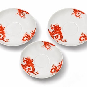 Könnte beinhalten: Drei weiße Keramikuntertassen mit einem leuchtend roten, verzierten Blumendesign. Jede Untertasse hat eine zentrale, vertiefte Fläche und ein sich wiederholendes Muster aus stilisierten Blättern und Wirbeln. Ein Set dekoratives Geschirr.