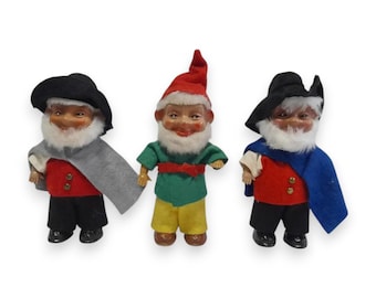 Puede incluir: Tres figuras de gnomos vintage. Cada gnomo tiene barba blanca, mejillas rosadas y una expresión sonriente. Visten diferentes atuendos: uno con chaleco rojo y sombrero negro, otro con camisa verde y pantalón amarillo, y el último con chaleco rojo y capa azul.