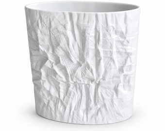 May include: A white ceramic vase with an oval opening and a textured exterior surface that mimics the appearance of crumpled paper. The vessel features deep folds and creases throughout its body, creating a complex pattern of light and shadow.