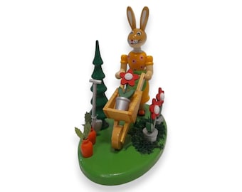 Puede incluir: Figura de madera de un conejo empujando una carretilla llena de flores. El conejo viste un vestido naranja con lunares amarillos. También hay un árbol verde, una pala y zanahorias. La figura está sobre una base verde.