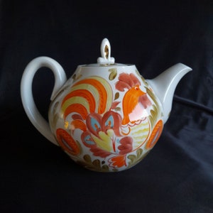 Könnte beinhalten: Weiße Porzellan-Teekanne mit einem Hahn-Design. Der Hahn ist in Orange, Gelb und Blau mit grünen Blättern bemalt. Die Teekanne hat einen goldenen Knopf auf dem Deckel.