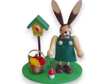 May include: A wooden Easter bunny figurine wearing a green apron, standing on a green base. The bunny has brown ears and is next to a birdhouse and a basket of flowers. A small mushroom is also on the base.