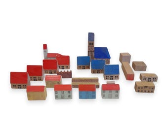 May include: A collection of wooden toy blocks shaped like buildings, including houses, a church, and other structures. The blocks are painted in various colors, such as red, blue, and beige, and are arranged on a white background.