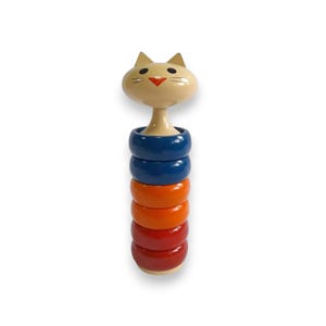 May include: A wooden stacking toy featuring a cat head on top of colorful rings. The cat head is beige with black eyes and a red nose. The rings are blue, orange, and red.