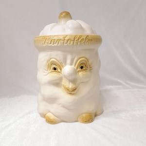 May include: A vintage ceramic cookie jar in the shape of a smiling face. The jar is primarily off-white with tan accents, including the rim and facial features. The word "Kartoffeln" is written around the rim.