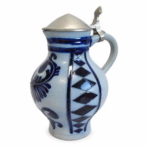 Op de afbeelding: Een lichtblauwe keramische bierpul met een metalen deksel en handvat. De buik is versierd met donkerblauw handgeschilderd bloemmotief en een verticaal paneel met donkerblauwe ruiten. Het deksel is conisch en voorzien van een scharnier.