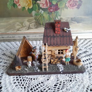 以下が含まれることがあります： 屋根が茶色で、煙突と小さな鳥が乗っている木製のミニチュアハウス。家の前に小さな人形が2体立っていて、1体は道具を持ち、もう1体はバスケットを持っている。家の前に小さな犬がいる。