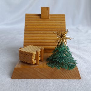 May include: A miniature wooden Christmas scene featuring a log cabin, a small green Christmas tree with a gold star topper, and a stack of logs. The scene is set on a wooden base, perfect for holiday decor.