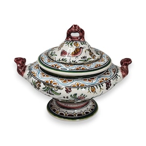 May include: A decorative ceramic lidded dish with a detailed floral and bird design. The dish features a white base with colorful accents of blue, green, and brown. It has two handles and a small pedestal base.
