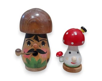 Puede incluir: Dos figuras de hongos de madera. Uno tiene una gorra marrón, una cara pintada con bigote y una pipa. El otro tiene una gorra roja con lunares blancos, un cuerpo blanco y una cara sonriente. Sobre un fondo blanco.