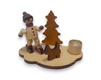 Puede incluir: Figura navideña de madera que representa a un niño patinando sobre hielo con un perro y un árbol de Navidad. La figura está pintada en tonos marrones y beige, con un pequeño portavelas. La base es de color marrón claro, con forma de nube.