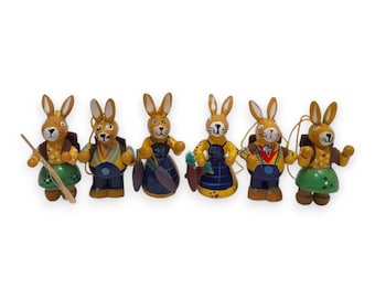 Peut inclure: Six figurines de lapins de Pâques en bois. Chaque lapin est peint avec une tenue et des accessoires uniques, notamment des tabliers, des sacs à dos et des carottes. Les lapins sont principalement bruns, avec des accents de bleu, de jaune et de vert.