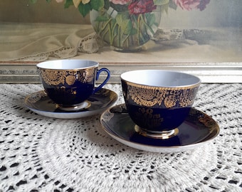 May include: Two blue and gold teacups and saucers with intricate floral designs. The cups and saucers are on a white crocheted tablecloth.