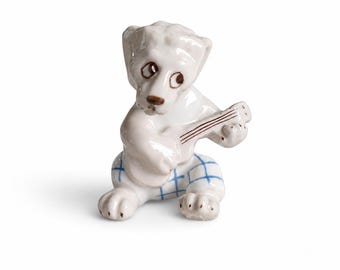 May include: A small white ceramic figurine of a dog sitting down and playing a guitar. The dog has brown eyes and a brown nose, and is wearing white pants with a blue grid pattern. The guitar is also white with thin brown painted strings.