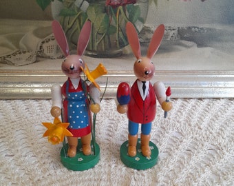 May include: Two wooden bunny figurines, one holding a painted egg and a paintbrush, the other holding a bouquet of yellow flowers. Both bunnies are wearing colorful clothing and standing on green bases.