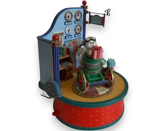 May include: A whimsical Christmas-themed music box featuring a bear character in a green vehicle, holding a gift. The box has a blue backdrop with clocks, a map, and a sign that reads "Post Office". The base is red with a dotted pattern.