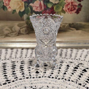 May include: A clear glass bud vase with a cut glass design and three legs. The vase is sitting on a white crocheted doily.