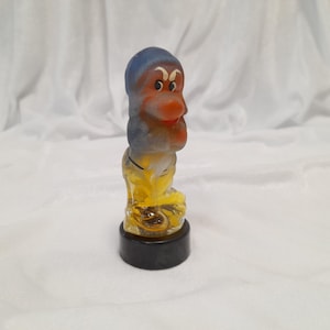 May include: A glass figurine of a dwarf from the Disney movie Snow White and the Seven Dwarfs. The dwarf is wearing a blue hat and yellow pants. The figurine is on a black base.