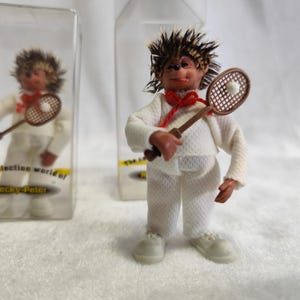 May include: A figurine of a hedgehog wearing a white tennis outfit, holding a brown racket with a white ball. The hedgehog has a red bow tie and spiky hair. The figurine is in a clear plastic case.