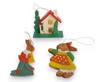 Puede incluir: Tres adornos navideños de madera. Uno es una casa con tejado rojo y base verde. Dos son figuras de conejos, una con un atuendo rojo y blanco, la otra con un vestido amarillo con lunares naranjas. Cada adorno tiene una cuerda plateada para colgar.