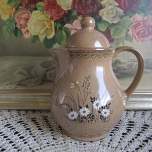Puede incluir: Una tetera de cerámica marrón con un diseño floral con flores blancas y hojas verdes. La tetera tiene una tapa y un asa.