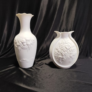 May include: Two white ceramic vases with floral relief designs. One vase is tall and slender with a ruffled rim, the other is a shorter, rounded vase. Both vases have detailed embossed floral patterns.