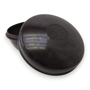May include: A dark brown, circular, lidded container with a smooth, glossy finish. The lid features a subtle, embossed circular logo in the center. The container is slightly open, revealing a hollow interior and a matching dark brown base.
