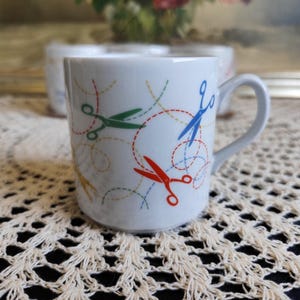 Könnte beinhalten: Weiße Keramiktasse mit grauem Henkel, verziert mit bunten Scheren und gepunkteten Linien. Die Scheren sind rot, grün und blau. Die Tasse steht auf einer weißen Spitzendecke.