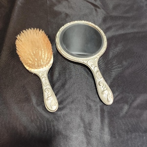 Op de afbeelding: Een vintage toilettafelset met een handspiegel en een borstel. Beide items hebben sierlijke zilverkleurige handvatten met een bloemmotief. De borstel heeft natuurlijk gekleurde borstelharen en de spiegel heeft een rond reflecterend oppervlak.