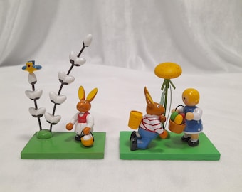 May include: Two wooden figurines of bunnies holding baskets of Easter eggs. One bunny is standing next to a white flower with a blue bird. The other bunny is kneeling next to a girl figurine holding a basket of eggs. Both figurines are on green bases.