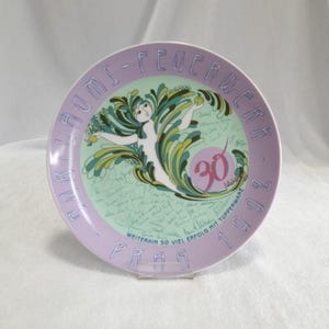 Könnte beinhalten: Ein dekorativer Teller mit lila Rand und einem zentralen Design mit einer stilisierten Figur mit fließenden grünen und blauen Akzenten. Der Teller hat Text am Rand und eine "30" in der Mitte.