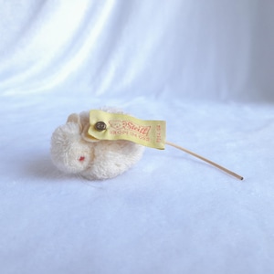 Puede incluir: Un ratón de peluche blanco con una etiqueta amarilla de Steiff que dice "Steiff Knopf im Ohr" y un número. El ratón está acostado de lado con la cola extendida.