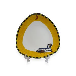 Può includere: Un piatto decorativo dal design stravagante. Il piatto presenta un centro bianco circondato da un bordo giallo con un bordo a scacchi nero e verde. Sul piatto è raffigurata una nave a vapore dei cartoni animati, insieme a una luna crescente.