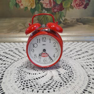 Puede incluir: Reloj despertador rojo con esfera blanca y agujas negras. El reloj tiene una campana a cada lado y la palabra "emco" impresa en la esfera.