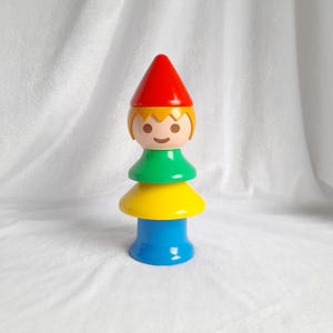 Puede incluir: Un juguete apilable colorido con forma de gnomo. El juguete presenta un sombrero cónico rojo, una cara con pelo amarillo, una sección media verde, una sección inferior amarilla y una base azul. Juguete para niños.
